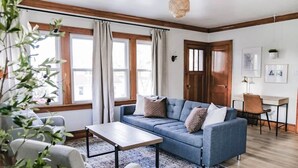 Un salon comprenant un canapé, une table basse et un bureau avec une chaise. On y trouve des fenêtres avec des rideaux, un luminaire suspendu et une plante fixée au mur.