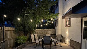 Une terrasse extérieure avec une table et des chaises, des guirlandes lumineuses, une clôture en bois et un arbre.
