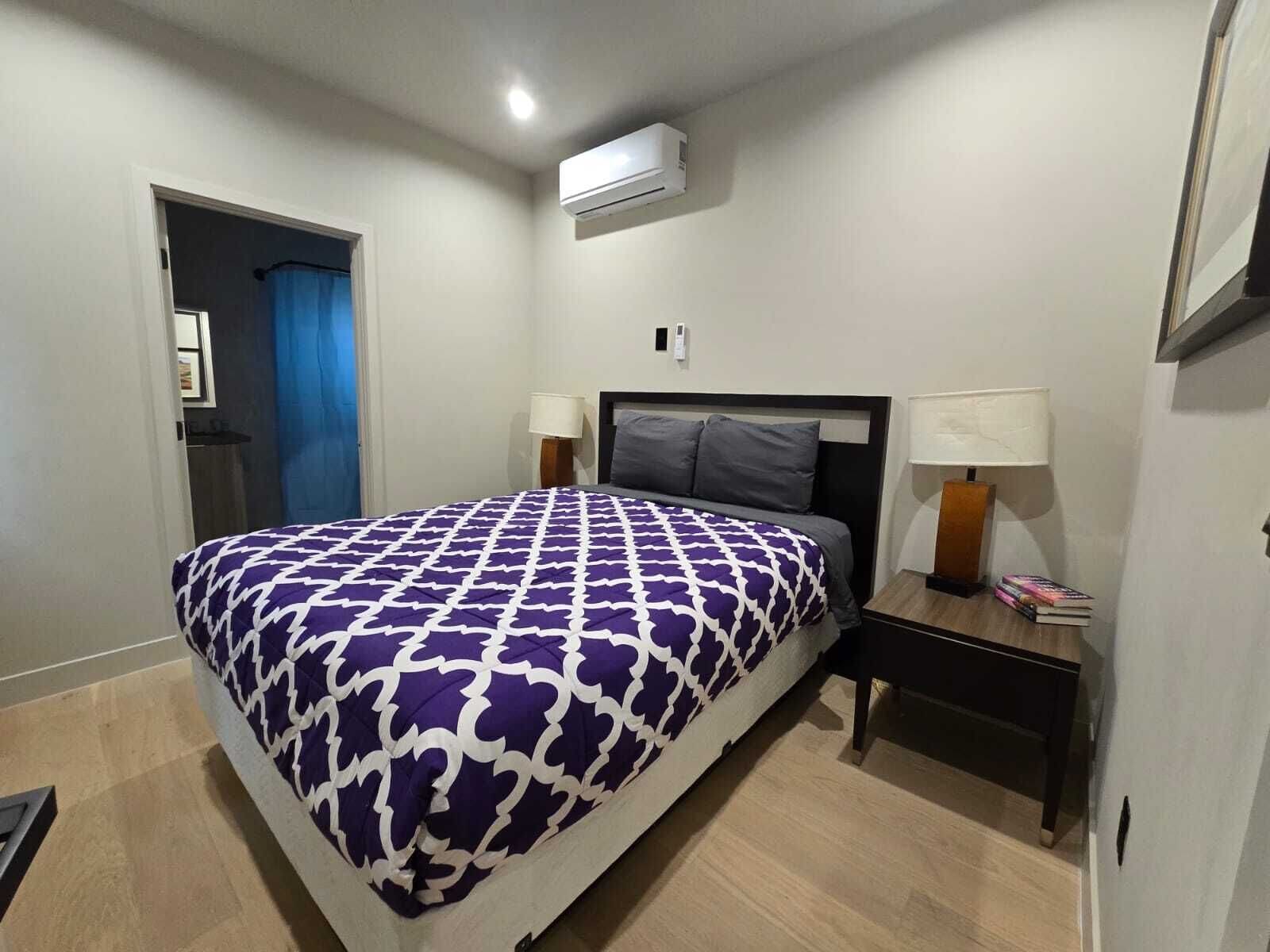 A hotel room with a bed, two bedside tables with lamps, a wall-mounted air conditioner, and a door with a blue curtain.