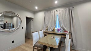Un coin repas agrémenté d’un miroir rond, d’une table avec des chaises et d’une vue sur la cuisine, visible à travers une fenêtre ornée de rideaux.