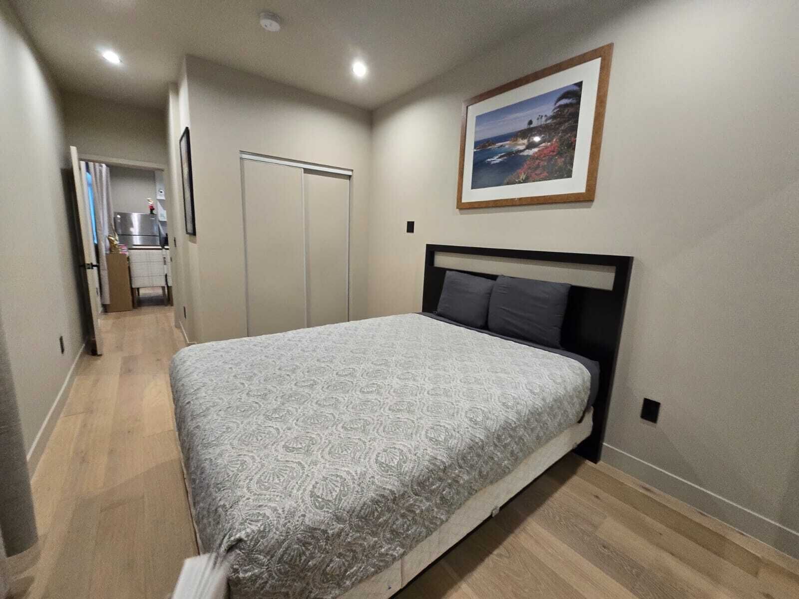 A bedroom with a bed, a wardrobe, and a kitchenette in the background.