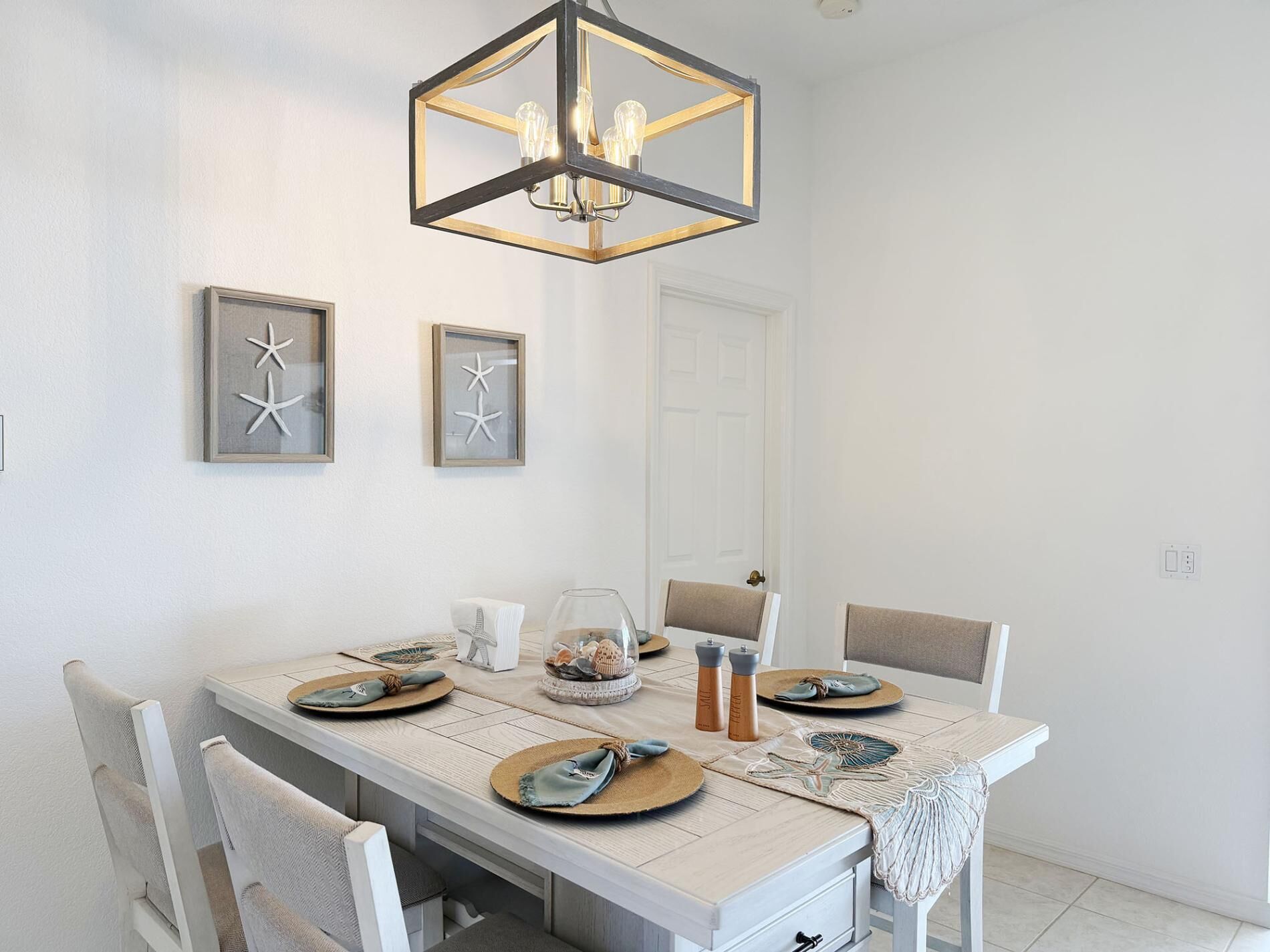 Uma sala de jantar com uma mesa retangular, cadeiras brancas, um candelabro e arte decorativa de estrelas-do-mar emoldurada na parede.