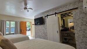 Habitación de hotel con dos camas, ventilador de techo, televisor y baño con espejo, además de una puerta que da al exterior.