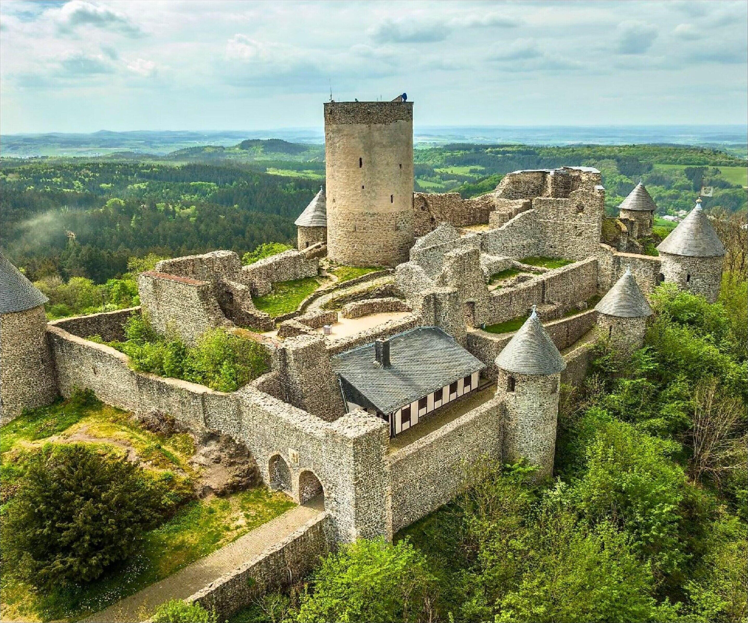 Une vue aérienne d’un château historique en pierre, doté de nombreuses tours et d’un donjon central, entouré d’une végétation luxuriante et de collines ondoyantes.