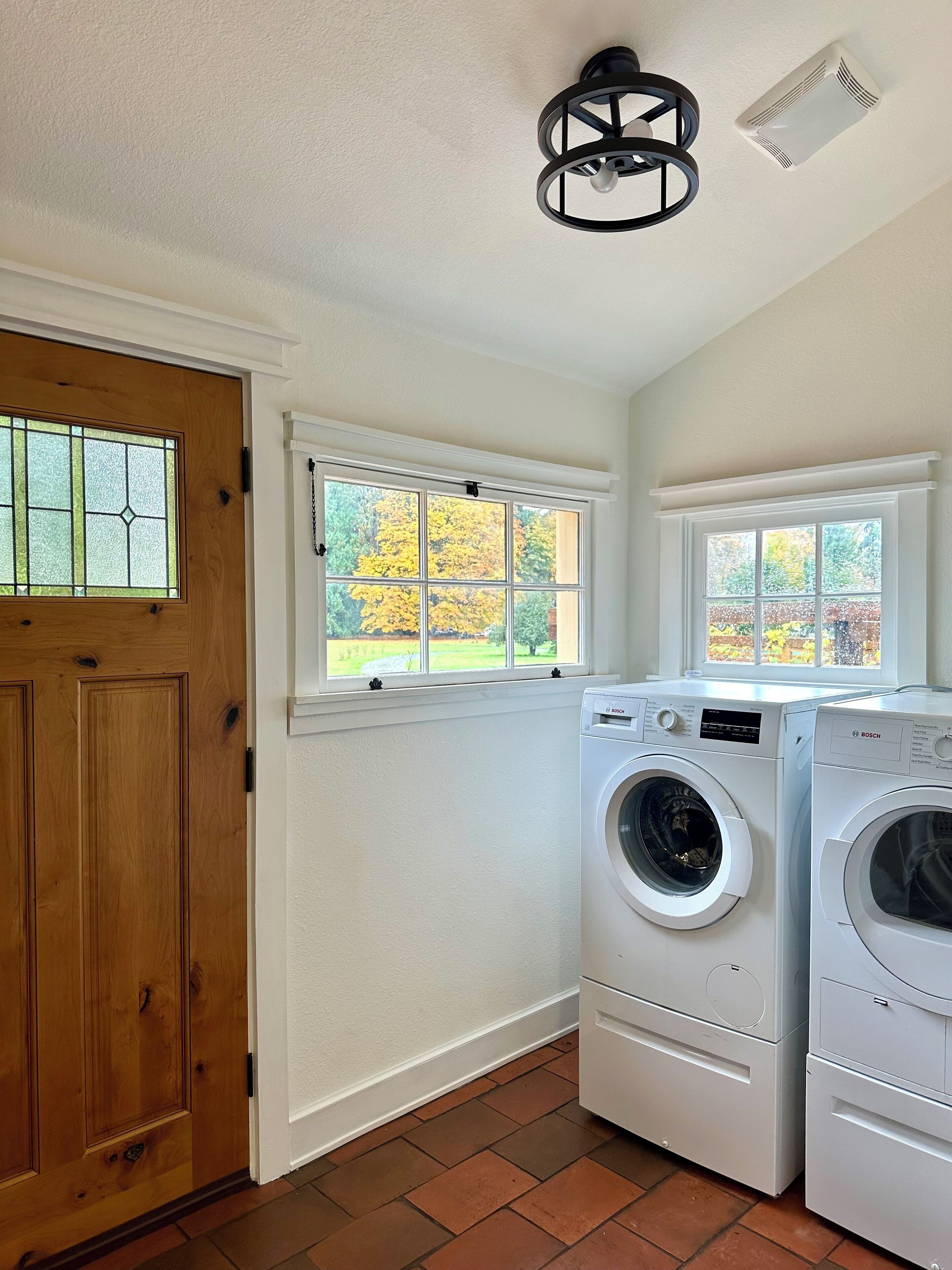 Un lavadero con dos lavadoras Bosch, una puerta de madera y ventanas que dan a árboles otoñales.