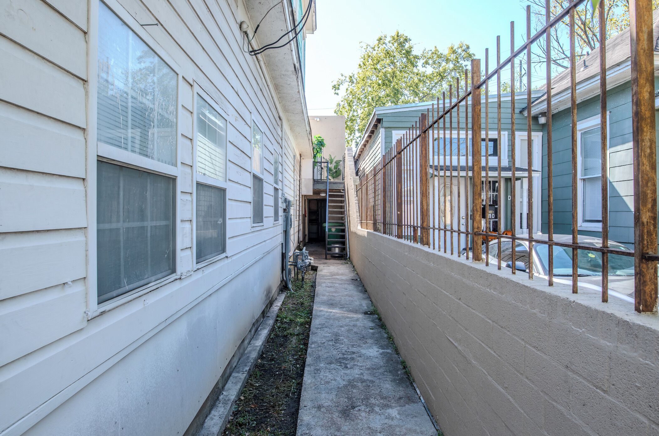A narrow alleyway between two buildings with a concrete path, a metal fence, and a staircase leading to a second level.