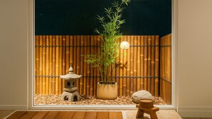 Una stanza con vista attraverso una finestra, che include un recinto in bambù, una lanterna di pietra, una pianta di bambù in vaso e un piccolo sgabello di legno.