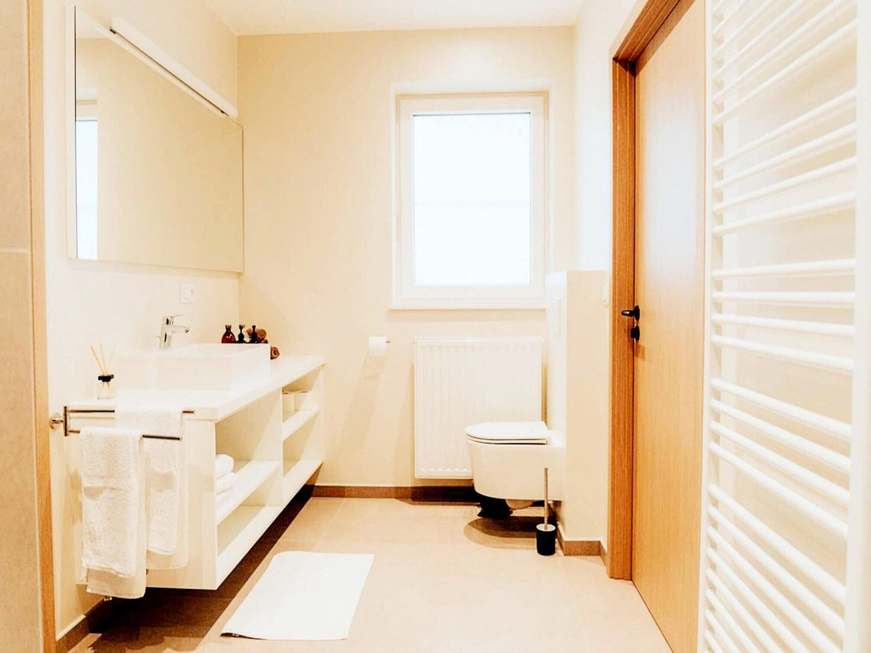 Un baño con lavamanos blanco, espejo y inodoro. Hay una ventana sobre el inodoro y un perchero con toallas.