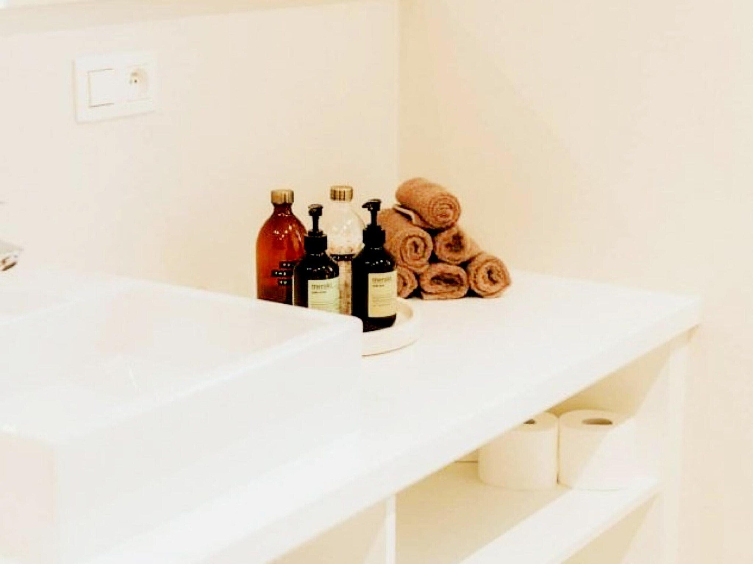 A bathroom with a white sink, toiletries, and rolled towels on a shelf.