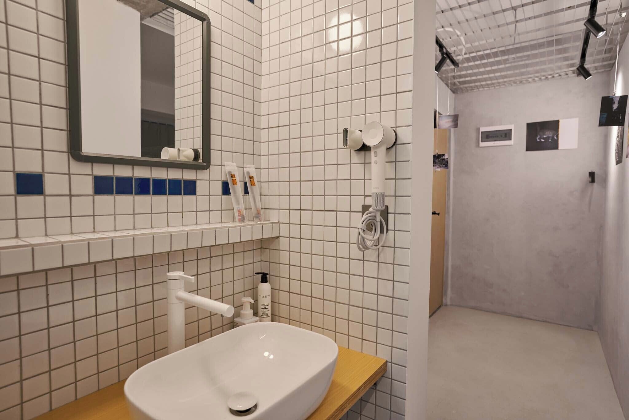 Un baño con lavamanos blanco, espejo y paredes con azulejos. Hay artículos de aseo en la encimera y se ve una zona de ducha al fondo.