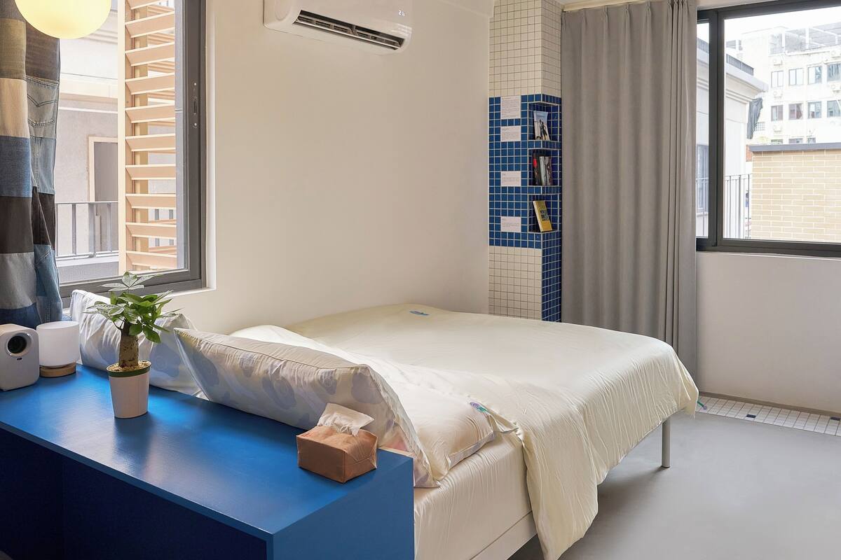 A hotel room with a bed, a blue bedside table with a potted plant, a white bedside table, and a window with curtains.
