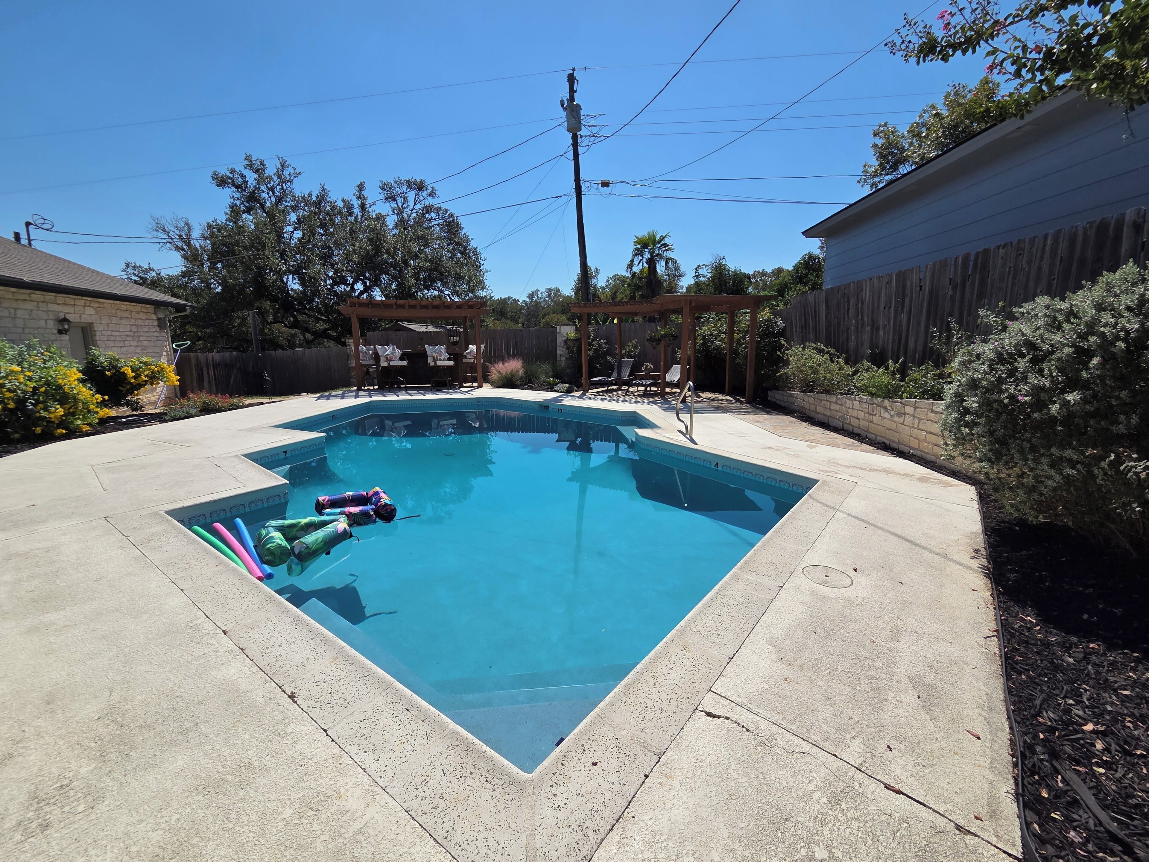 A rectangular swimming pool with a wooden deck surrounding it, a pergola with chairs, and a wooden fence in the background.