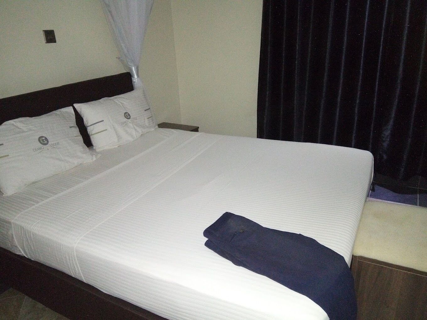 A neatly made bed with white linens and a dark blue blanket, a wooden headboard, and a small bedside table.