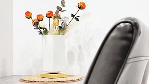 Un vase blanc orné de fleurs, posé sur une table, un tableau encadré accroché au mur et une chaise noire.