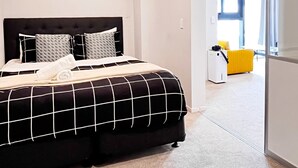 Une chambre moderne avec un grand lit, une table de chevet, une armoire et une vue sur une pièce lumineuse où se trouve un fauteuil jaune.