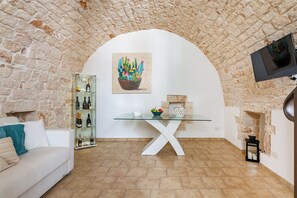 Un salon doté d’une voûte en pierre, d’une table en verre avec un pied en forme de X blanc, d’un canapé blanc, d’une vitrine contenant des bouteilles de vin et d’un tableau représentant un cactus.