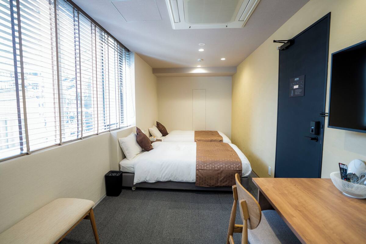 Quarto de hotel com cama, escrivaninha, cadeira, banco e janela com persianas.