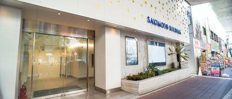 A building with a glass entrance, a sign reading "SAKIMOTO BUILDING," and a planter with plants outside.