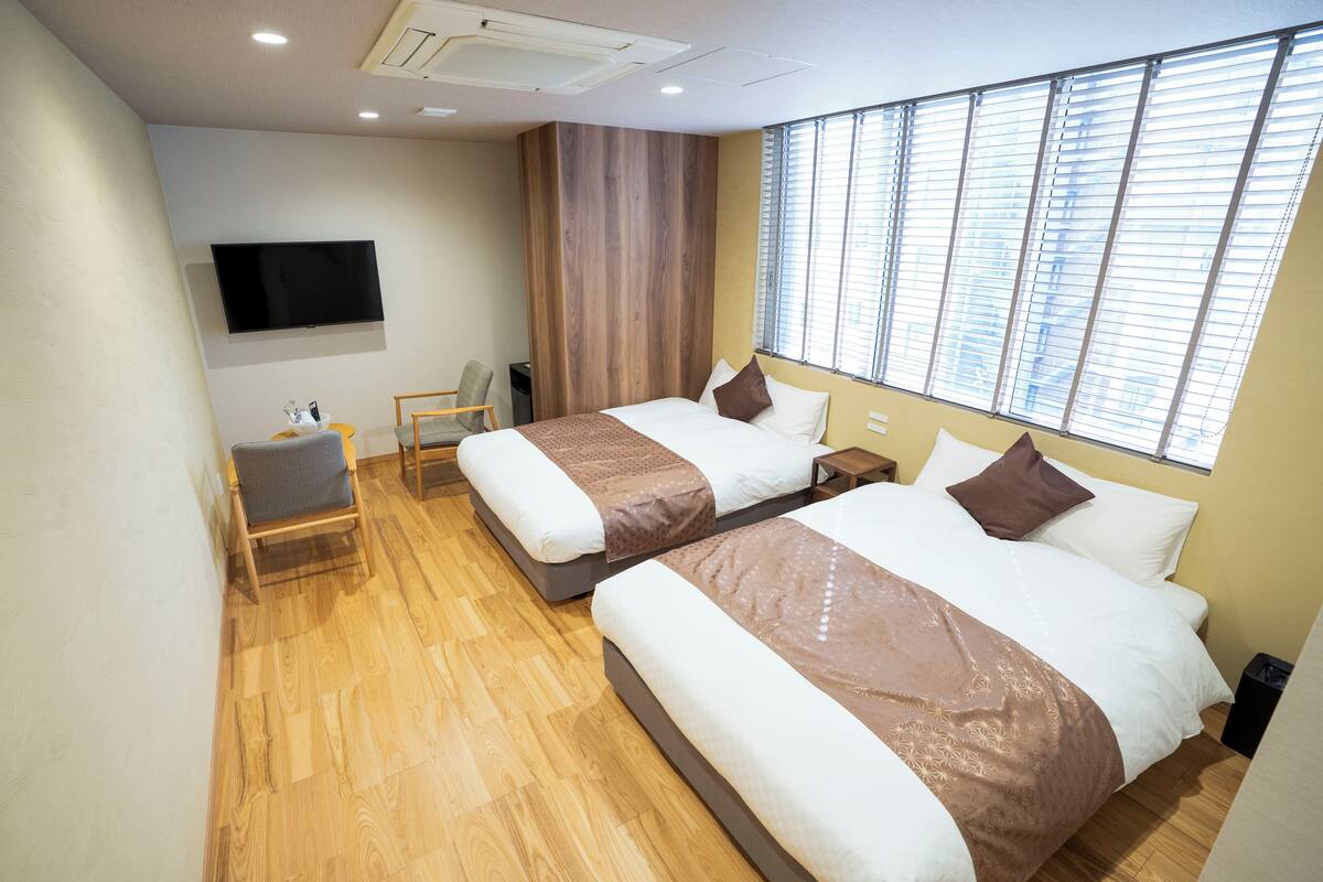 Quarto de hotel com duas camas, televisão, cadeira e janela com persianas.