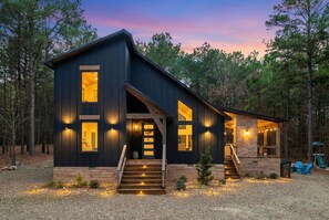 A modern two-story cabin with a dark exterior, large windows, and a wooden staircase leading to the entrance. - Hot Tub, Firepit, Arcade, Playset & Outdoor Lounge (Broken Bow)
