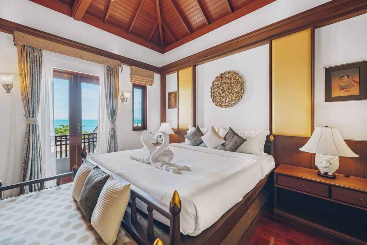 A hotel room with a large bed, a nightstand, a lamp, a wall decoration, and a view of the sea through a balcony.