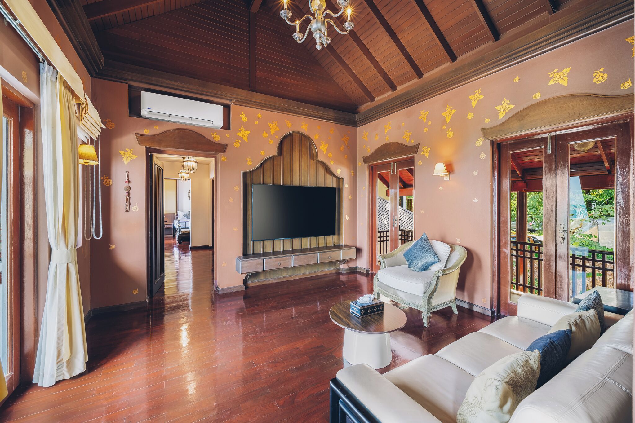 A spacious living room with a large flat-screen TV, wooden flooring, and a chandelier.