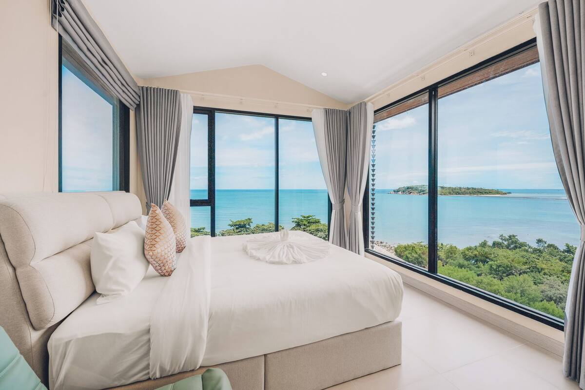 A bedroom with a large bed, two windows, and a view of the sea.
