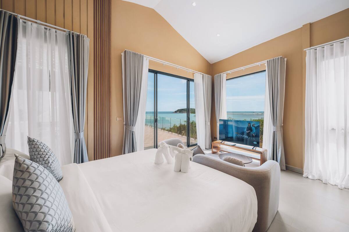 A modern hotel room with a large bed, a seating area, and a TV. The room has large windows offering a view of the sea.