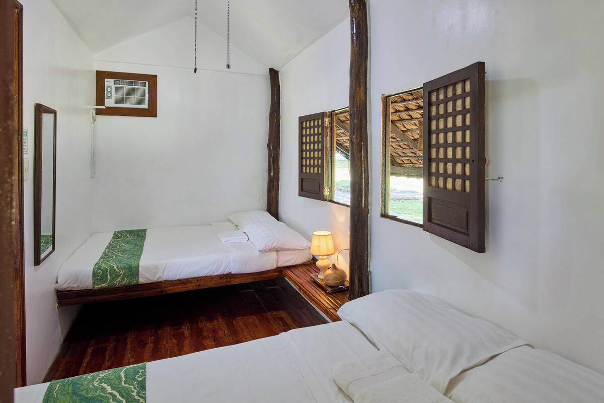 A small, single-room accommodation with a wooden bed, a bedside table with a lamp, and a window with wooden shutters.