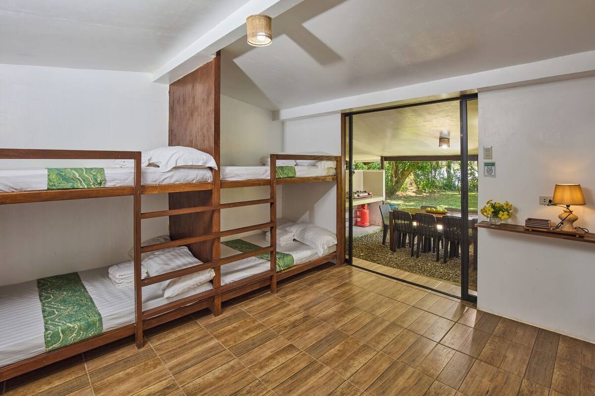 A dormitory room with bunk beds, a small kitchenette, and a view of an outdoor garden.