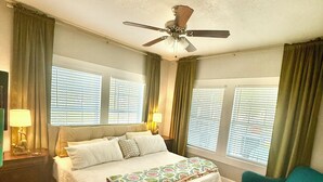 Un dormitorio con cama, ventilador de techo, dos ventanas con persianas, un sillón verde y una alfombra.