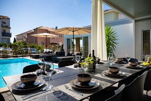 Area esterna con zona pranzo e piscina, tavoli apparecchiati con piatti, ciotole e bicchieri, e un grande ombrellone che offre ombra.