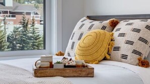 A bedroom with a large window, a bed with patterned pillows, a tray with items, and a view of buildings and trees. - Stunning Modern 2BR Unit in Downtown Canmore! (Canmore)