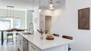 A modern kitchen with a central island, pendant lights, and a view of the adjacent dining area. - Stunning Modern 2BR Unit in Downtown Canmore! (Canmore)