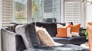 A modern living room with a dark grey sofa, multiple cushions, and a view of mountains through large windows. - Stunning Modern 2BR Unit in Downtown Canmore! (Canmore)