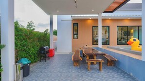 Een terras buiten met een houten tafel en banken, een barbecue en een vuilnisbak.