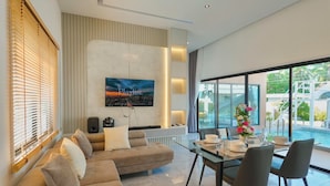 A modern living room with a sectional sofa, a dining table with chairs, a mounted TV, and a view of an outdoor swimming pool. - Pool&Play - Modern Private Pool Villa in Pattaya (Muang Pattaya)