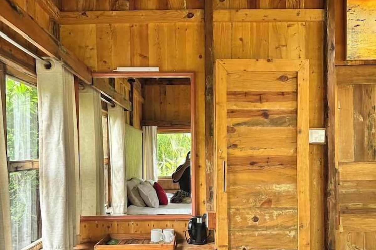 Eine Holzhütte mit einem Bett, einem kleinen Tisch mit Tassen und einer Kaffeemaschine sowie einem Fenster mit Vorhängen.