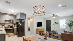 A modern kitchen with stainless steel appliances, a dining area with wooden chairs and placemats, and a living room with a sofa and a ceiling fan.