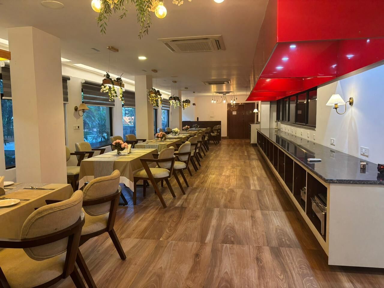 A restaurant with long tables, chairs, hanging plants, and a kitchen area.