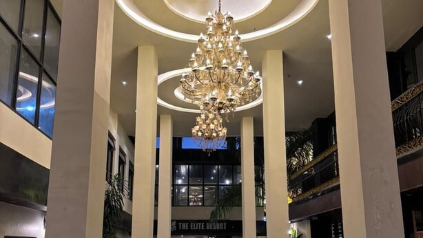 Ein geräumiger Innenhof mit einem großen Chandelier, hohen Decken und einer zentralen Säule, die zum Eingang eines Hotels mit der Aufschrift „The Elite Resort“ führt.