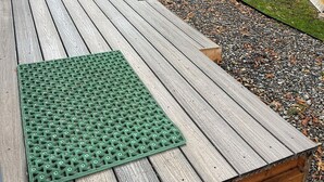 Un rivestimento in legno con un tappetino verde, circondato da un giardino con aiuole rialzate e una recinzione in legno.