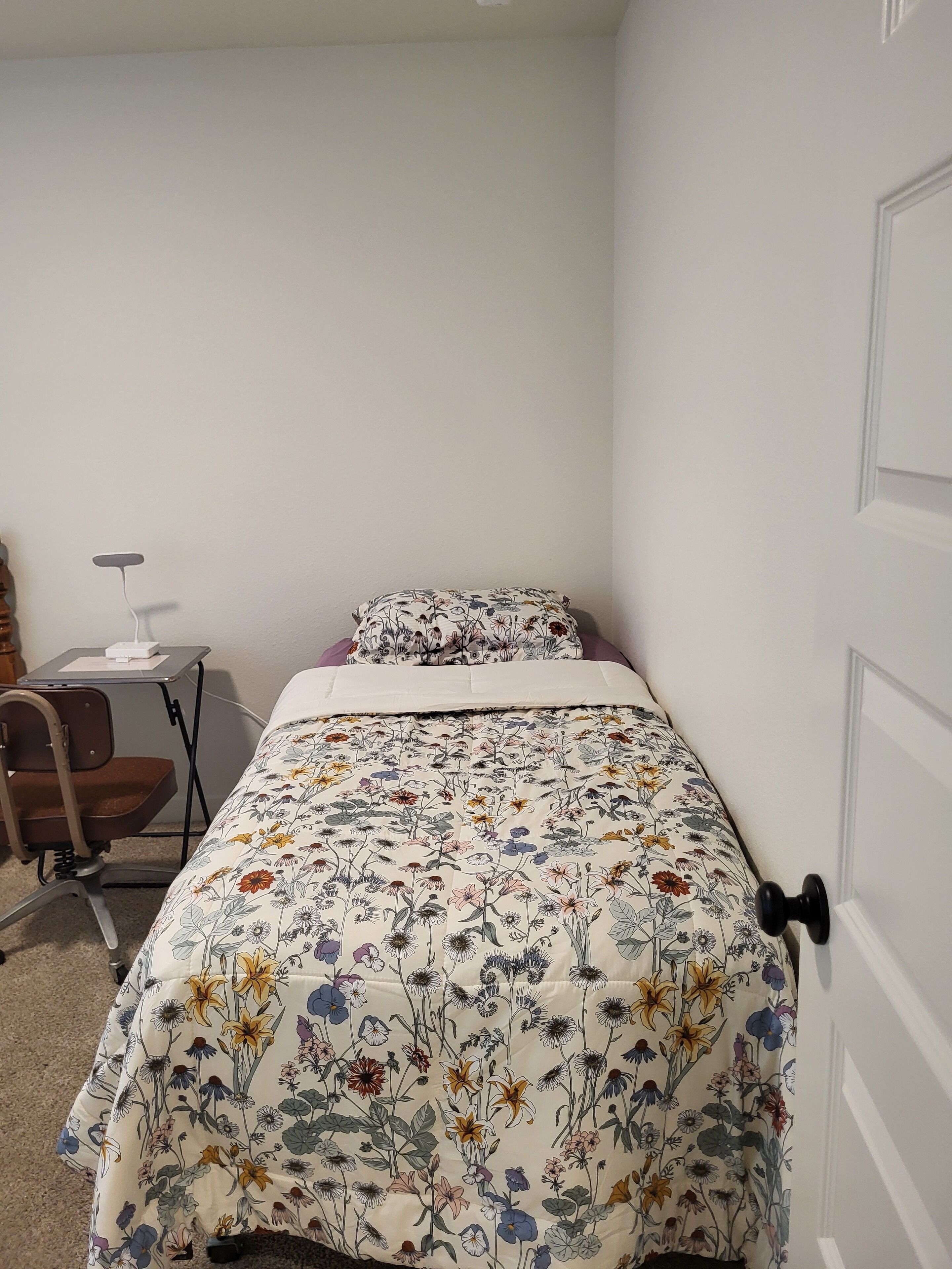 A small, neatly arranged bedroom with a bed, a chair, a desk, and a door.