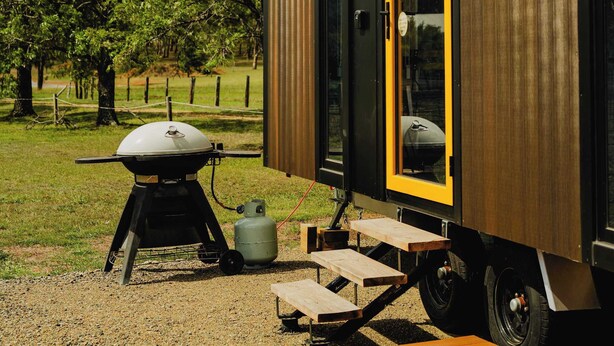 A modern, compact mobile home with a large window and a door, a barbecue grill, and a picnic table outside. - Sunset Tiny House at Bluebush Estate X by Tiny Away (Rothbury)
