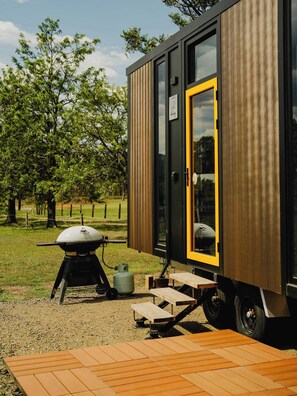 A modern, compact mobile home with a large window and a door, a barbecue grill, and a picnic table outside. - Sunset Tiny House at Bluebush Estate X by Tiny Away (Rothbury)