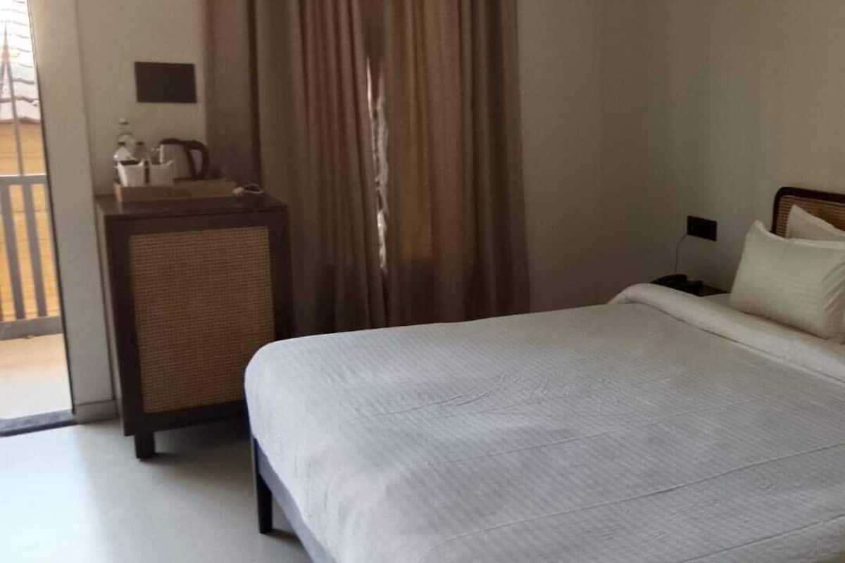 A hotel room with a bed, a nightstand, a lamp, a small table with a pitcher, and a view of the outside.