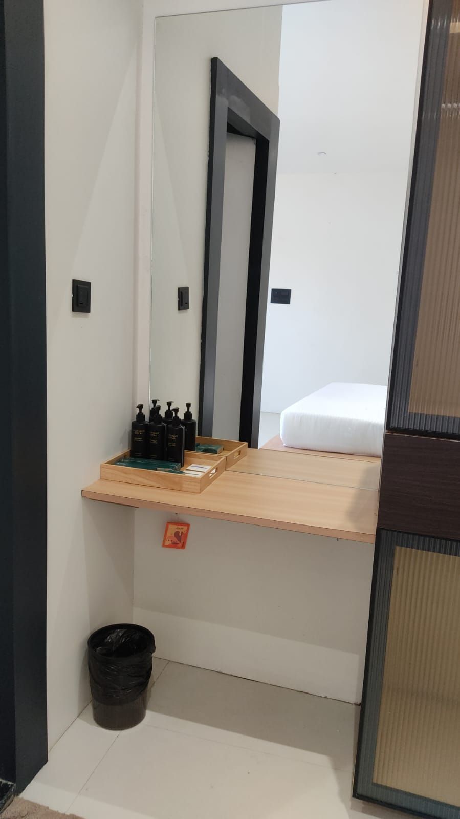 A small, compact room with a wooden shelf holding toiletries, a single bed, a mirror, and a trash bin.