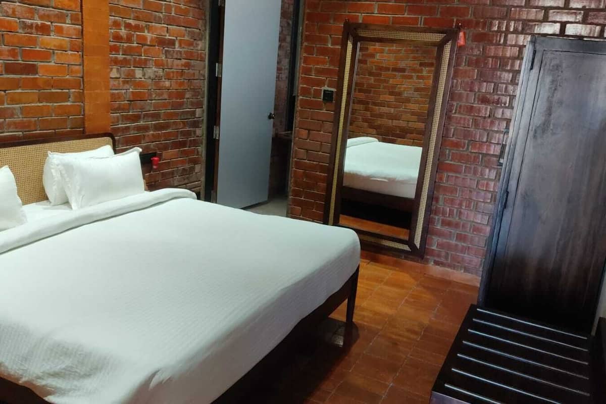 A hotel room with a brick wall, a bed with white linens, a wardrobe, and a mirror.