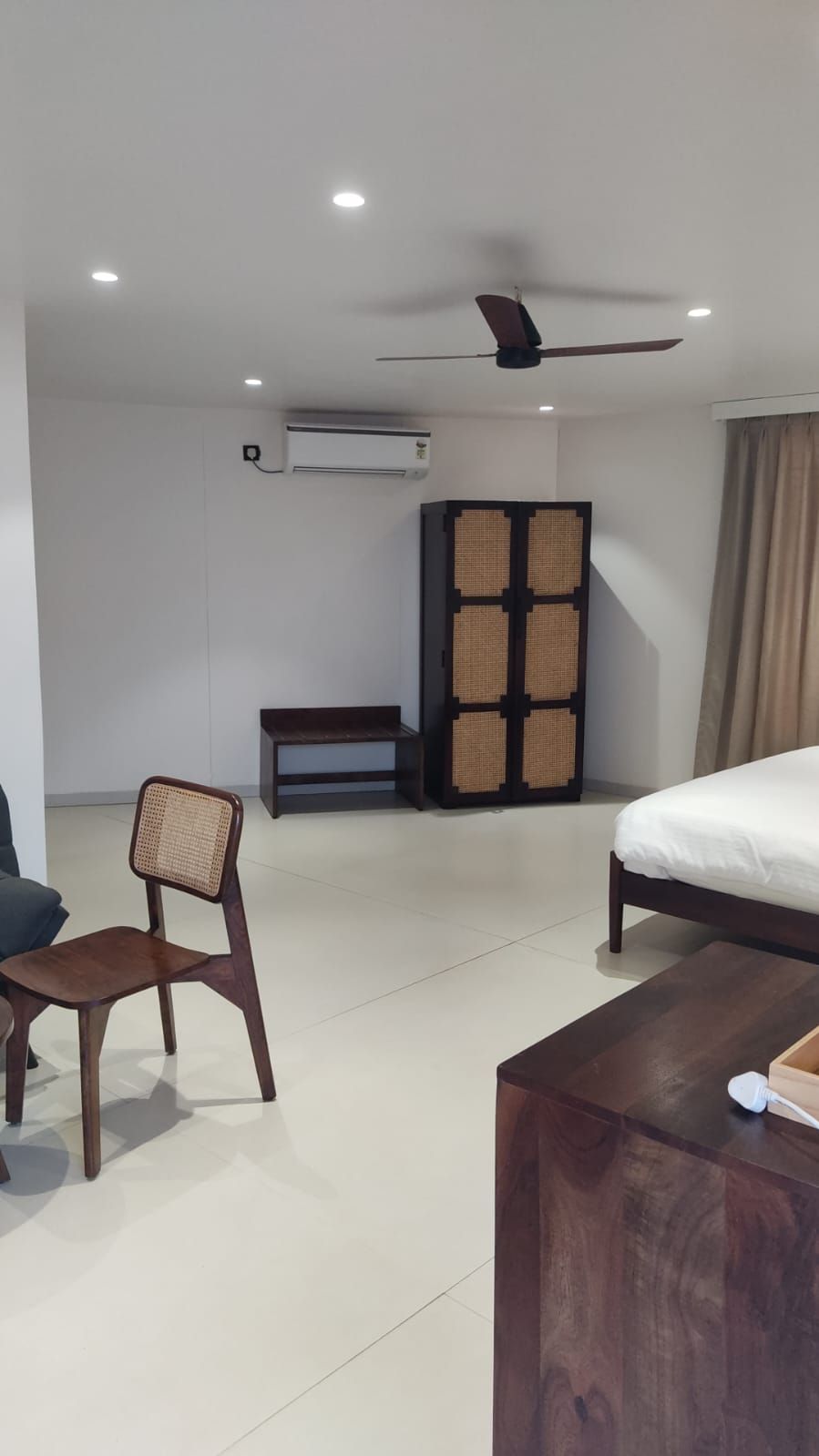 A hotel room with a bed, a chair, a bench, a wardrobe, a ceiling fan, and an air conditioning unit.