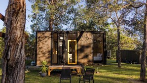 Une petite cabane avec une porte, un banc, deux chaises et un foyer à l’extérieur.